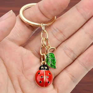 May include: A gold-toned metal keychain featuring a round split ring attached to a short chain. The chain holds two charms: a round, red enamel ladybug with black spots and a small, green enamel leaf charm with detailed black vein markings.