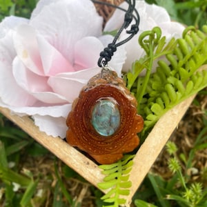 May include: A pendant necklace featuring a polished, oval-shaped iridescent blue stone set within a natural, textured brown wood slice. The pendant hangs from a thin, black cord with an adjustable knot mechanism, showcasing organic wood grain.