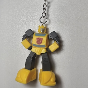 May include: A yellow and dark gray robot keychain featuring a character with a red Autobot insignia on its chest. The figure has a blocky design with yellow limbs and torso, dark gray joints, and a silver face with blue eyes, attached to a metal ring.