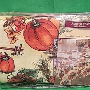 May include: An oblong fabric tablecloth labeled Autumn Fest measuring 60 in. x 102 in. The tablecloth features a cream background with a print of orange pumpkins, green leaves, wheat stalks, and a brown wooden fence. The packaging shows a table setting.