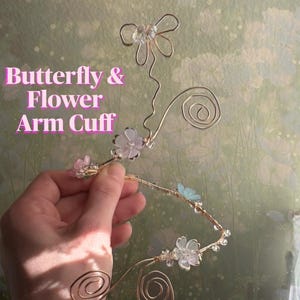 May include: A gold-toned wire arm cuff featuring a butterfly shape at the top, spiral ends, and small translucent pink, white, and blue flower accents. The text "Butterfly & Flower Arm Cuff" is printed in pink on the left side of the image.