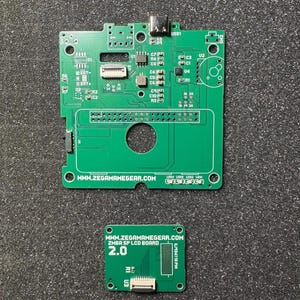 Puede incluir: Una placa de circuito impreso verde con un puerto USB-C, varios componentes electrónicos y el texto WWW.ZEGAMAMEGEAR.COM, junto a una placa LCD verde más pequeña etiquetada WWW.ZEGAMAMEGEAR.COM ZMBA SP LCD BOARD 2.0 con un conector de cable plano.