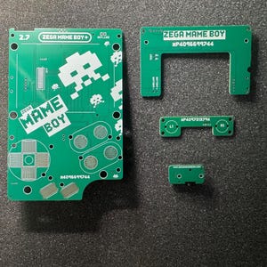 Puede incluir: Un conjunto de cuatro placas de circuito impreso verdes para un proyecto Zega Mame Boy. La placa más grande presenta gráficos de alienígenas pixelados en blanco, el texto Zega Mame Boy y contornos de botones. Las placas pequeñas incluyen etiquetas L1, R1.