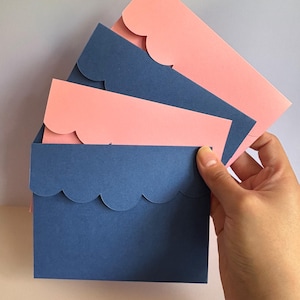 May include: A set of four rectangular envelopes featuring scalloped edges on the flaps. The envelopes alternate in color between solid matte pink and solid matte navy blue, stacked in a fan-like arrangement with the blue envelope in the front.