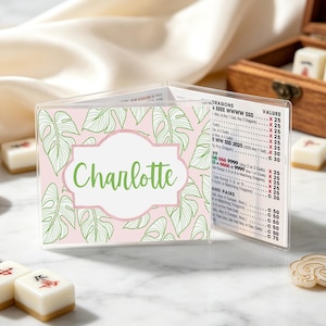 May include: A clear plastic Mahjong score card holder featuring a light pink background with green monstera leaf line art. The center label displays the name Charlotte in green script. The right side shows a printed Mahjong scoring guide.