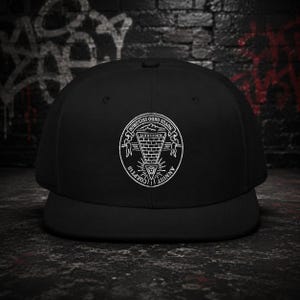 May include: A solid black baseball cap featuring a white circular graphic on the front. The design depicts an inverted pyramid with an eye at the centre, surrounded by the text ANNUIT COEPTIS and NOVUS ORDO SECLORUM with MDCCLXXVI at the top.