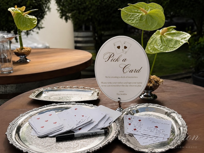 K&ouml;nnte beinhalten: Ein rundes wei&szlig;es Schild mit dem Text Pick a Card, We're creating a deck of memories, Please write your wishes and sign your name so we can remember this day when we play, und The Smiths, ausgestellt mit Spielkarten auf silbernen Tabletts.