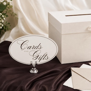 May include: A rectangular white card box with a thin ribbon bow and a slot on top, accompanied by an oval sign on a silver stand featuring the dark brown text "Cards AND Gifts" in a script font, alongside a light beige envelope on a dark fabric.