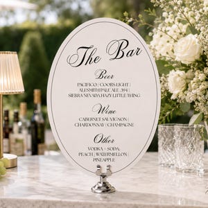 Op de afbeelding: Een ovaal wit barmenu-bord met zwarte tekst die The Bar, Beer, Wine en Other leest, met een lijst van diverse drankopties. Het bord wordt rechtop gehouden door een zilveren metalen standaard op een marmeren oppervlak.