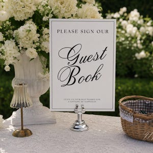 May include: A rectangular white guest book sign with black text that reads PLEASE SIGN OUR Guest Book and LEAVE US YOUR BEST WISHES FOR A LIFETIME OF HAPPINESS. The sign is held upright by a small silver metal stand on a white lace tablecloth.