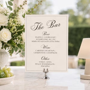 May include: A rectangular white bar menu sign with black text reading The Bar, Beer, Wine, and Other, listing various drink options. The sign is held by a silver metal stand, positioned next to a white floral arrangement and a small white lamp.