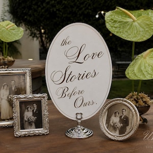 May include: An oval white sign with gold cursive text reading the Love Stories Before Ours, supported by a silver stand, accompanied by three silver-toned picture frames containing black and white wedding photographs and green anthurium flowers.