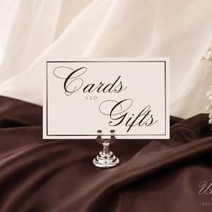 May include: A rectangular off-white sign with a thin dark brown border and the text "Cards AND Gifts" in a dark brown script font. The sign is held upright by a small, polished silver metal stand with a circular base and a clip top.