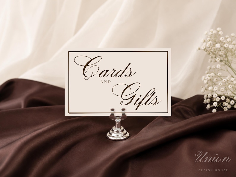 May include: A rectangular off-white sign with a thin dark brown border and the text "Cards AND Gifts" in a dark brown script font. The sign is held upright by a small, polished silver metal stand with a circular base and a clip top.