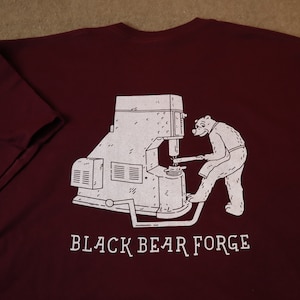 Puede incluir: Camiseta burdeos con una ilustración blanca de un oso en una forja. El oso está operando una máquina grande. El texto "BLACK BEAR FORGE" está impreso debajo de la ilustración.