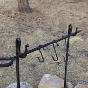 May include: Black metal cooking rack with multiple hooks and a ring on the side. The rack is standing on two metal legs.