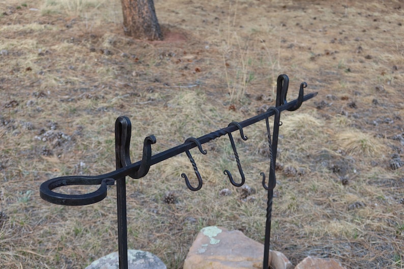 May include: Black metal cooking rack with multiple hooks and a ring on the side. The rack is standing on two metal legs.
