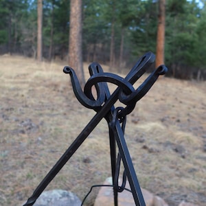May include: A black metal tripod stand with a unique, decorative top. The stand is designed to hold a pot or kettle over a fire.