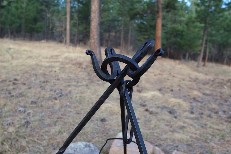 May include: A black metal tripod stand with a unique, decorative top. The stand is designed to hold a pot or kettle over a fire.