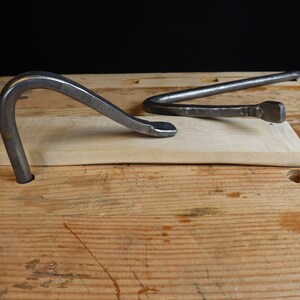 May include: Two hand-forged metal tools rest on a light-colored wooden plank. One tool is a curved, hook-like shape, while the other is a bent bar with a squared end. The tools are set against a dark background on a wooden surface.