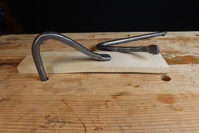 May include: Two hand-forged metal tools rest on a light-colored wooden plank. One tool is a curved, hook-like shape, while the other is a bent bar with a squared end. The tools are set against a dark background on a wooden surface.