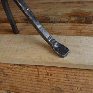 May include: Close-up of a metal tool with a flat, beveled end resting on a light-colored wooden plank. The tool is dark gray and appears to be hand-forged. The wood plank is rectangular and sits on a wooden surface.