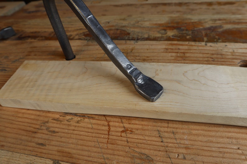 May include: Close-up of a metal tool with a flat, beveled end resting on a light-colored wooden plank. The tool is dark gray and appears to be hand-forged. The wood plank is rectangular and sits on a wooden surface.