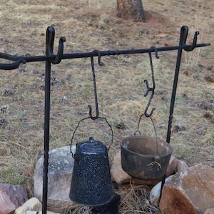 May include: A black metal tripod stand with hooks for hanging pots and kettles over a fire. A blue enamel pot and a black cast iron pot are hanging from the hooks.