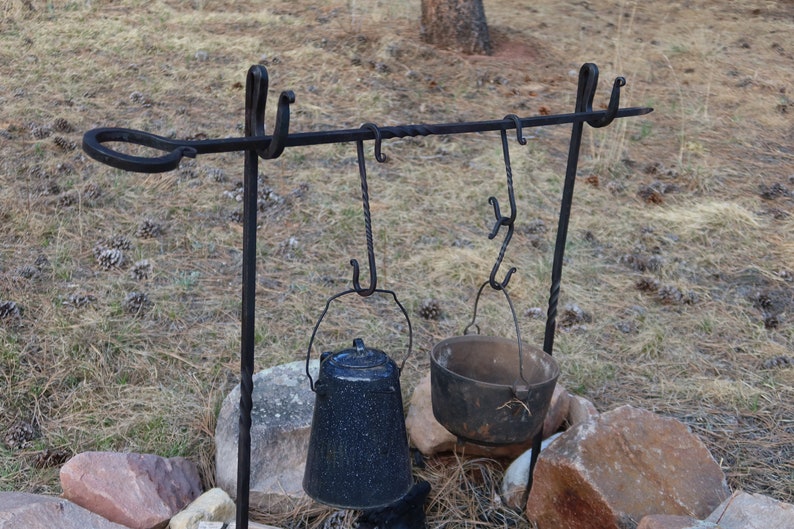 May include: A black metal tripod stand with hooks for hanging pots and kettles over a fire. A blue enamel pot and a black cast iron pot are hanging from the hooks.