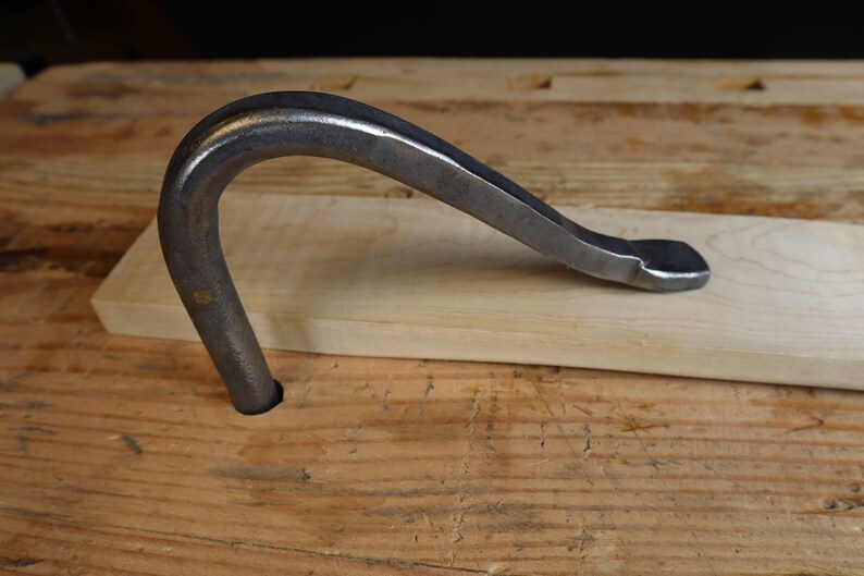 May include: A metal woodworking tool with a curved handle and a flat, beveled end. The tool is dark gray and rests on a light-colored wooden plank. The background is a wooden surface.