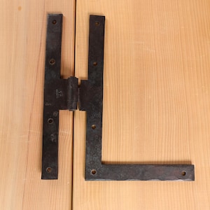 Puede incluir: Una bisagra en forma de H de hierro forjado negro con un acabado rústico. La bisagra está montada sobre una superficie de madera clara. Tiene múltiples orificios para tornillos para una fijación segura.