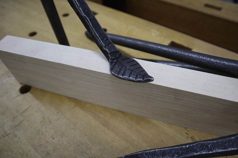 Pu&ograve; includere: Primo piano di uno strumento in metallo nero a forma di foglia che riposa su una superficie in legno chiaro. Lo strumento fa parte di un set di strumenti utilizzati per la lavorazione del legno.