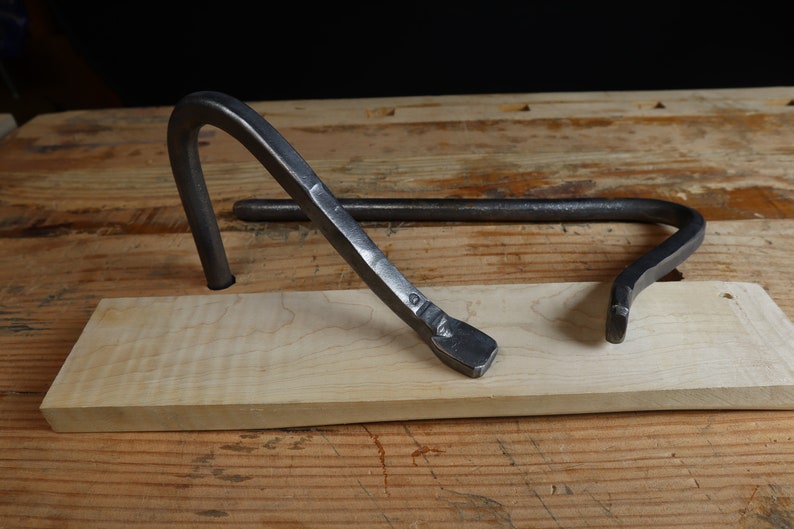 May include: A hand-forged metal tool with a curved design, resting on a light-colored wooden block. The tool has a dark, textured finish and appears to be a specialized pry bar or lever. The background is a wooden surface.