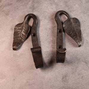 May include: Two handcrafted, dark metal hooks with a leaf design. Each hook features a curved top, a leaf-shaped accent, and a rectangular base. The hooks are likely for hanging items and have a rustic, vintage aesthetic.