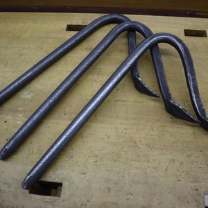 Pu&ograve; includere: Tre ganci in metallo nero con forma curva e estremit&agrave; appuntite. I ganci sono disposti in fila su una superficie di legno.