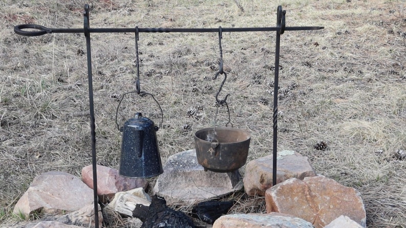 May include: A black metal tripod stand with a horizontal bar and two hooks. A black kettle and a cast iron pot hang from the hooks over a fire pit.