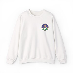 May include: A white long-sleeve crewneck sweatshirt featuring a circular graphic on the left chest. The design depicts a stylized mountain range in green and white under a dark blue sky with a golden sun, all enclosed within a thin circular border.