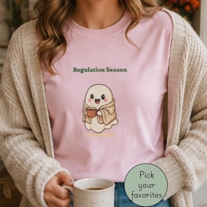 May include: A light pink t-shirt featuring a graphic of a smiling ghost wrapped in a tan blanket and holding a brown mug. Above the ghost, the text reads Regulation Season in dark green. The shirt is worn under a beige textured cardigan.