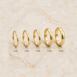 May include: Five polished gold rings arranged in a row, increasing in width from left to right. The rings are labeled with their respective widths of 2 mm, 3 mm, 4 mm, 5 mm, and 6 mm, indicating the specific thickness of each gold band.