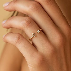 May include: A gold-toned open ring featuring a capital letter D on one end and a single small clear round gemstone on the other end. The band is thin and metallic, designed to be worn on a finger with the letter and stone facing outward.