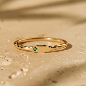 Può includere: Un sottile anello a fascia in oro con una superficie liscia e lucida, caratterizzato da una piccola pietra preziosa circolare di un verde vibrante incastonata a filo nel metallo. L'anello ha un profilo delicato e arrotondato.