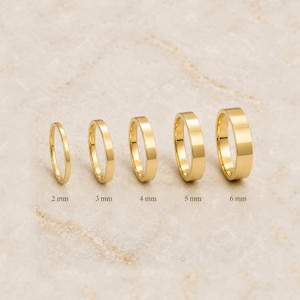 Può includere: Cinque anelli color oro disposti in fila dal più piccolo al più grande. Gli anelli sono etichettati con le rispettive larghezze di 2 mm, 3 mm, 4 mm, 5 mm e 6 mm. Ogni anello presenta una superficie liscia e lucida e un design a fascia piatta.