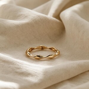 Puede incluir: Un anillo de oro con un diseño de banda continua y ondulada. La superficie metálica es lisa y pulida, reflejando la luz a lo largo de sus bordes curvos y rítmicos. El anillo mantiene una forma circular consistente con un patrón repetitivo.