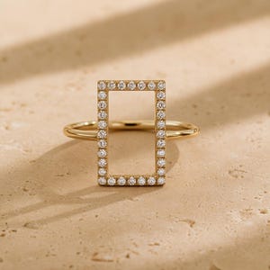 Op de afbeelding: Een gouden ring met een verticaal rechthoekig frame dat langs de gehele omtrek is bezet met kleine, heldere, rondgeslepen stenen. De ringband is glad en gepolijst en verbonden met de zijkanten van het rechthoekige middendeel.