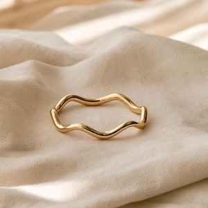 May include: A polished gold-tone ring featuring a continuous, smooth, undulating wave pattern. The metal band has a reflective surface and a consistent thickness throughout its circular shape, creating a fluid, rhythmic design that encircles the finger.