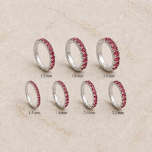 Könnte beinhalten: Sieben silberne Eternity-Ringe mit leuchtend roten Steinen, angeordnet in zwei Reihen. Die Ringe sind mit ihren jeweiligen Breiten beschriftet: 1,5 mm, 1,8 mm, 2,0 mm, 2,2 mm, 2,5 mm, 2,8 mm und 3,0 mm, was eine Auswahl an Ringgrößen zeigt.