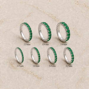 May include: Seven silver-toned eternity bands set with small green stones, arranged in two rows. Each ring is labeled with its width: 1.5 mm, 1.8 mm, 2.0 mm, 2.2 mm, 2.5 mm, 2.8 mm, and 3.0 mm. The rings feature a consistent circular design.