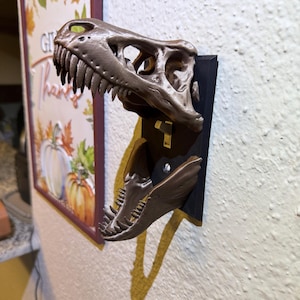 May include: A metallic bronze-colored Tyrannosaurus rex skull light switch cover plate mounted on a black rectangular base. The dinosaur skull is split into two parts, with the upper jaw forming the top and the lower jaw forming the bottom.