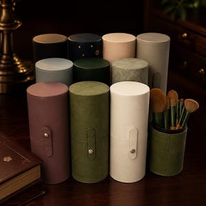 May include: A collection of cylindrical makeup brush holders in various colors including mauve, olive green, cream, black, navy blue, and gray. Each holder features a snap-button closure strap, and one open container displays a set of makeup brushes.