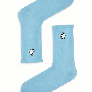 Pode incluir: Um par de meias de cano médio em azul claro com textura canelada, apresentando um pequeno gráfico bordado de um pinguim em preto e branco no lado exterior de cada meia. O design é simples, de cor sólida e com textura uniforme.